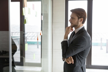 Pensive young Caucasian businessman look at glass wall in office think of project implementation, thoughtful serious male employee brainstorm develop business plan in boardroom, work with sticky notes