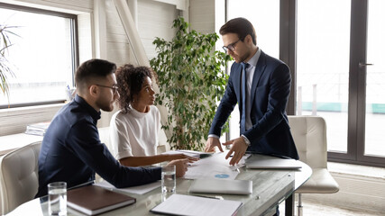 Concentrated multiracial businesspeople gather at desk in boardroom brainstorm over financial paperwork, diverse international colleagues talk discuss business project at meeting in office together