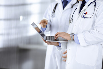 Two unknown doctors with stethoscopes discuss medical exam resoults, while standing at hospital office. Physicians using a laptop for checking up medication names records. Perfect medical service in