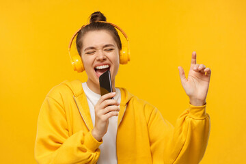 Young girl in yellow hoodie and wireless headphones, singing with closed eyes, holding smartphone...