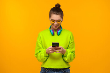 Teen girl wearing sweatshirt, colored eyeglasses and headphones around neck, looking at screen of phone with smile, isolated on yellow background
