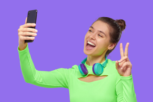 Young happy girl taking selfie photo with her phone, smiling and showing V sign, isolated on purple backgroound