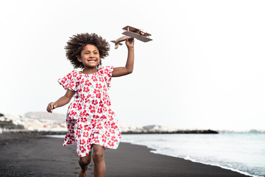 Afro child playing with wood toy airplane on the beach - Little kid having fun during summer holidays - Childhood and travel vacation concept