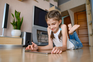 A girl in a white T-shirt lies on the floor. Girl playing on a tablet and smiling.