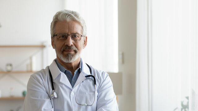 Headshot portrait of serious mature male doctor or therapist in white medical uniform, glasses and stethoscope, mature old man GP or physician in spectacles pose at workplace, healthcare concept