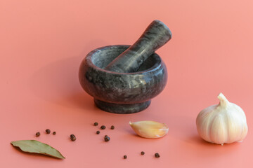 A marble mortar and pestle next to an artistically scattered whole black pepper, a Bay leaf and a garlic bulb on a pink background. Spices.