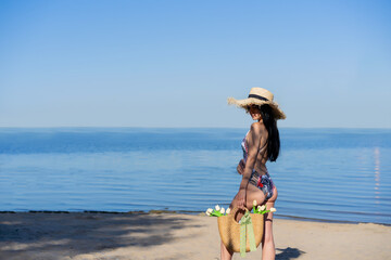 dark-haired beautiful girl in a swimsuit walks along the beach in a straw hat and with a wicker bag with flowers
