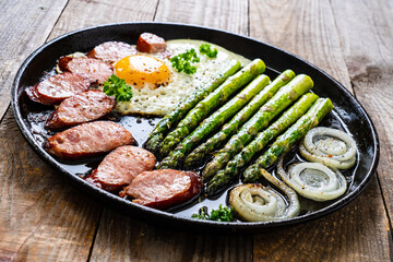 One pen breakfast. Sunny side up eggs with green asparagus, fried sausages and onion in pan on wooden table
