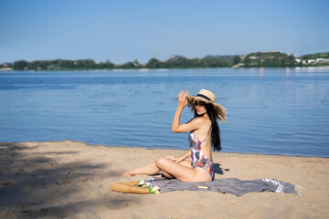 dark-haired beautiful girl in a swimsuit sitting on the beach in a straw hat
