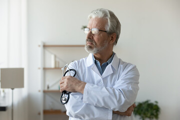 Pensive elderly male doctor or GP in glasses and medical uniform stand hold stethoscope look in...