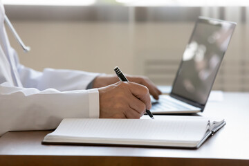 Crop close up of male doctor in white uniform sit at desk work on laptop fill patient anamnesis in medical journal, man GP or physician make notes in paper register, use computer, healthcare concept