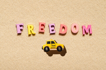 Toy yellow taxi car ride on the colorful letters of word freedom on beach sand. Concept. Fast and...