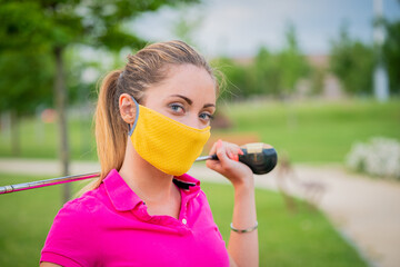 Portrait of a beautiful woman who playing golf on a background of green field outdoors with protective masks - Young girl with golf club stick - Close up blonde girl who is playing during quarantine