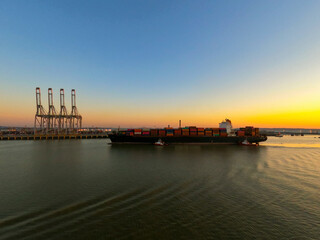 Big container ship vessel mooring in the port on the river during sunrise in calm weather / Container terminal and gantry cranes on back view