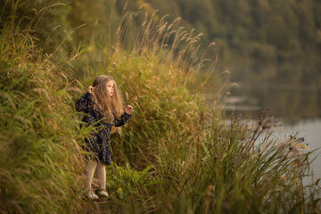 A small long-haired girl is standing in a thicket of grass on the Bank of the river. Image with selective focus.
