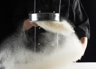 a dark background baker's hands sift white wheat flour over a sieve over