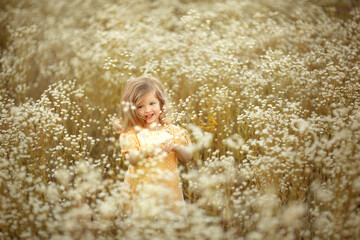 A little blonde girl is having fun and jumping in a field with small white flowers daisies. Image with selective focus.