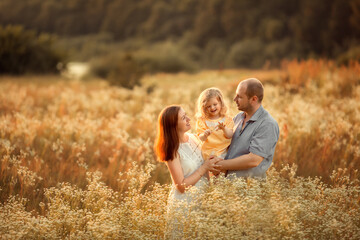 Naklejka na ściany i meble Mom, dad and their little daughter are having fun in a chamomile field near the forest. They are hugging and laughing. Image with selective focus.