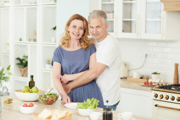 Waist up portrait of loving mature couple embracing and looking at camera while posing in white kitchen interior at home, copy space