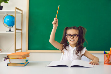 Back to school concept. Little girl schoolgirl with glasses raised her finger up has an idea on the background of a geenboard in elementary school.