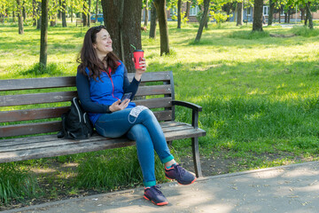 Obraz premium Beautiful Caucasian woman with long brown hair sitting in the park wearing a protective mask drinking a cup of tea or coffee and smiling. toned
