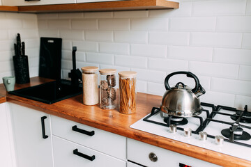 Glass jars in kitchen. Metallic teapot on the gas oven. Bright kitchen interior. White modern dining room. Wooden complete kitchen with gas oven.