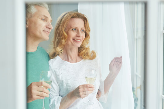 Waist Up Portrait Of Elegant Mature Couple Enjoying Champagne While Looking Out Of Window, Shot From Behind Glass, Copy Space