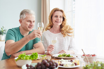 Portrait of loving mature couple raising champagne glasses while enjoying romantic dinner at home, copy space
