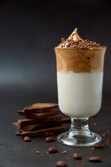 Glass of trendy fluffy creamy dalgona or whipped instant coffee with milk, coffee beans and chocolate on the dark background. New popular food and drink trend concept. Copy space.