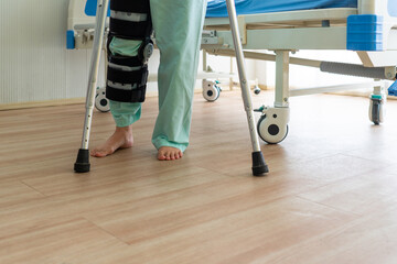 woman who wearing knee brace practices walking by using adjustable walking crutches after surgery...