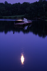 The moon is reflected off a placid lake with mountains & trees in the background and a swimming float in the mid-ground of the lake.