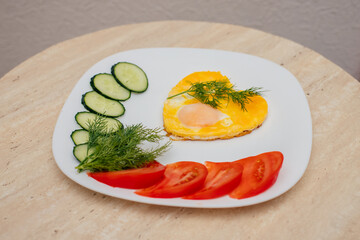 fried eggs in the form of a heart lie on a white plate, next to chopped dill, tomatoes and cucumbers