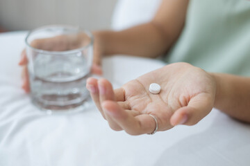 Adult woman holding pill and glass of water, taking medicine on bed in morning at home. Migraine, painkiller, headache, influenza, illness, sickness and healthcare concept
