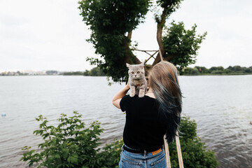 A girl with blue hair stands with her back and holds a three month old Scottish Straight kitten outside in the summer near a tree. The cat looks at the camera.. High quality photo