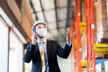 Asian warehouse manager in safety helmet wearing a medical surgical mask talking on the phone