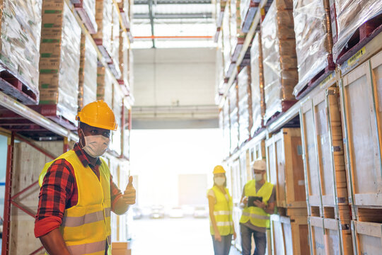 Warehouse Workers Team Meeting Inspect The Product Using A Tablet. With Wearing Face Mask And Protective Safety Helmets Standing In Storage Factory