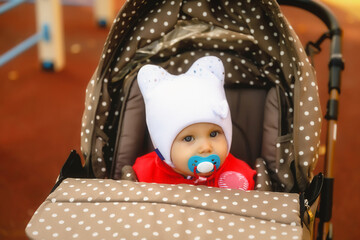Fototapeta na wymiar Baby with pacifier in its mouth in stroller. Cute happy blue-eyed one-year-old girl in red coat and white hat. Charming child shows emotions in close-up. Concept of healthy child and parenting