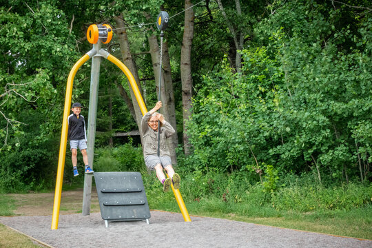Mature woman with glasses riding a zip-line at an outdoor playground at a public park. Child standing on the side looking.