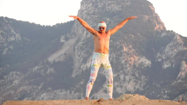 Young bald slender freak man tight leggings, topless, bandage on his head, remains with naked torso, backdrop of mountain, jumping, waving, depicting bird. Humor, unusual people. Slow motion video