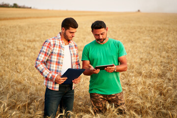 Two farmers stand in wheat stubble field, discuss harvest, crops. Senior agronomist with touch tablet pc teaches young coworker. Innovative tech. Precision farming with online data management soft.