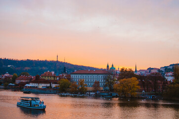 Naklejka premium Dramatic sundown in the Vltava River with a boat passing by and a gradient of orange to pink colors reflecting from the sky into the water.