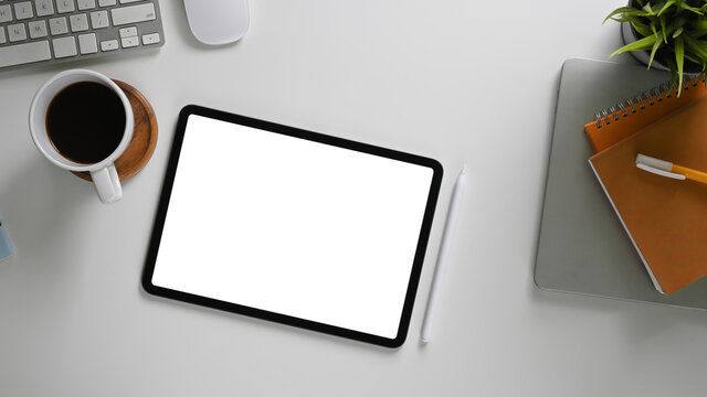 The top view image of a white blank screen computer tablet is putting on a comfortable working desk.