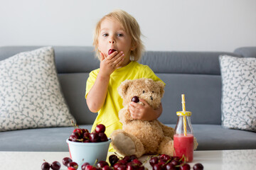 Toddler blond child, cute boy, eating cherries with teddy bears