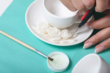 Roses with petals made of white polymer clay. A woman sticks them on a saucer. Corrects with a clerical knife. Crafts from polymer clay.