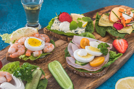 Variety of danish open sandwiches on wooden boards and blue background, decorated with vegetables, strawberries and aquavit