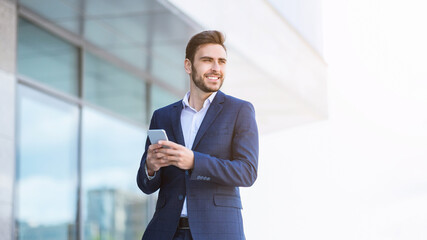 Smiling millennial industrialist checking his smartphone near skyscraper on city street