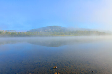Early foggy morning over the river - beautiful summer landscape. Thick fog over the water surface, reflection of hill with forest covered on the bank - freshness, calmness and enjoyment of nature