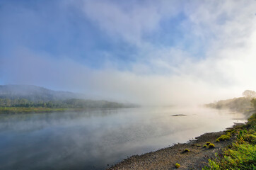 Early foggy morning over the river - beautiful summer landscape. Thick fog over the water surface, reflection of hill with forest covered on the bank - freshness, calmness and enjoyment of nature