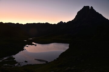 Le soleil se lève dans les montagnes françaises des Pyrénées, à côté de la frontière espagnole. Le pic du Midi d'Ossau se dresse au-dessus du joli lac de Miey. Le soleil est bleu, jaune et violet.