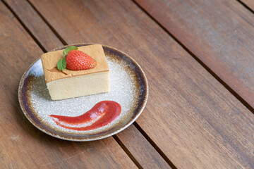 Japanese Cheesecake was turned into square pieces decorated with strawberry, placed on a ceramic plate that matches the brown wood floor.